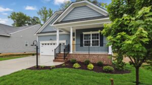 A blue Somerset Hill suburban house with a front porch, garage, green lawn, small shrubs, and a tree in the yard.