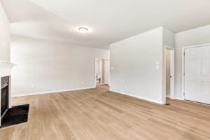 Bright, empty living room in Poplar Village with light wood floors, white walls, fireplace, and multiple doorways.
