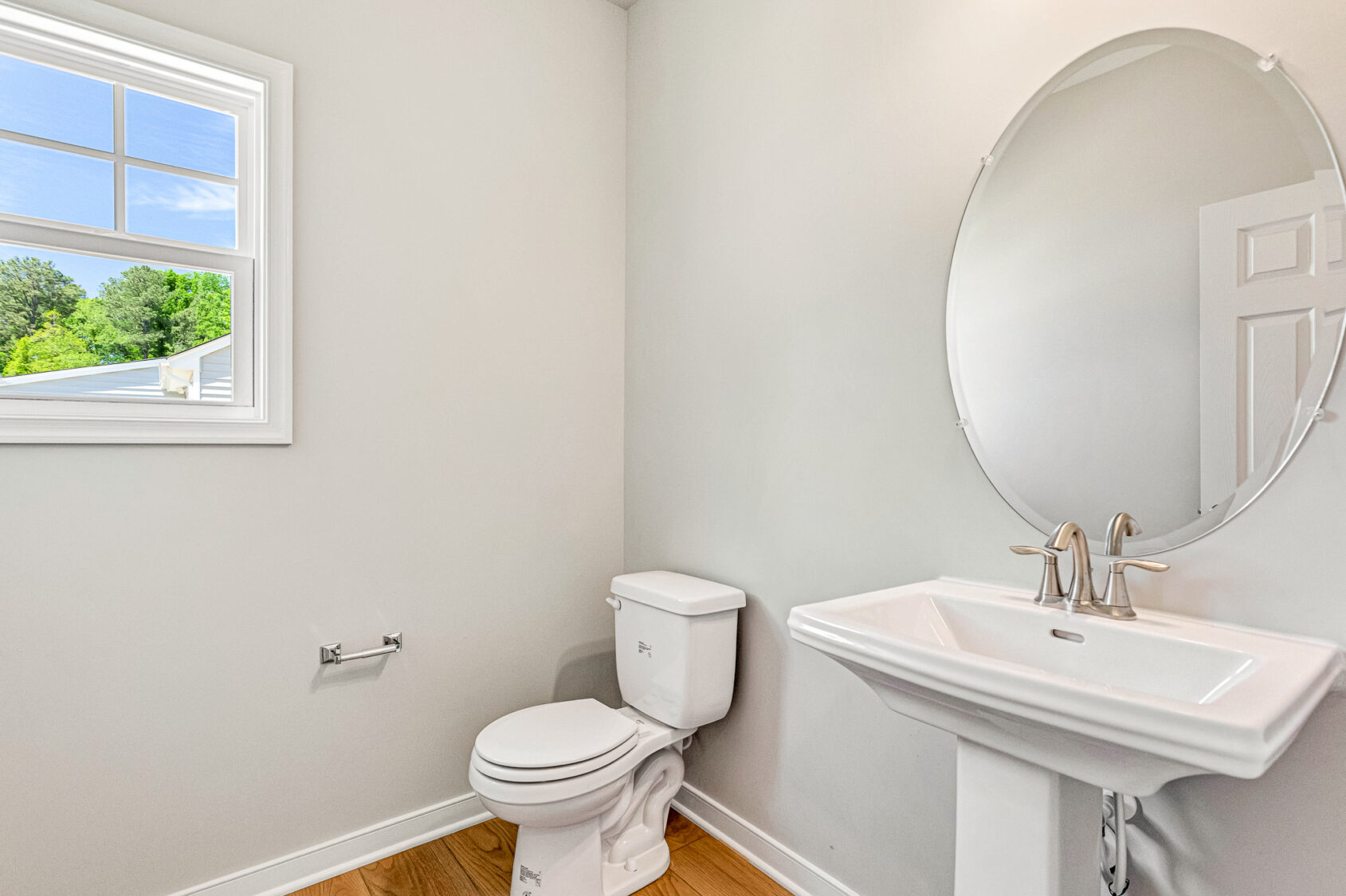 Bright bathroom in Poplar Village with a toilet, pedestal sink, oval mirror, hardwood floor, and window overlooking Lot 26.