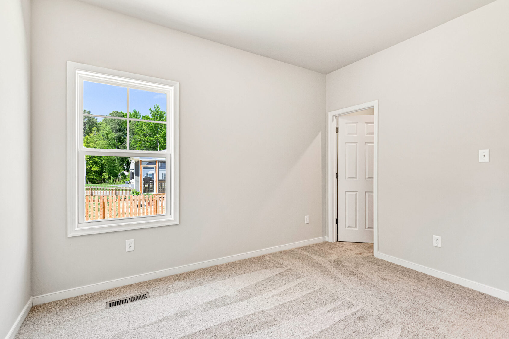 Bright, empty room in Poplar Village with beige carpet, white walls, window, and door to another area—Lot 26.