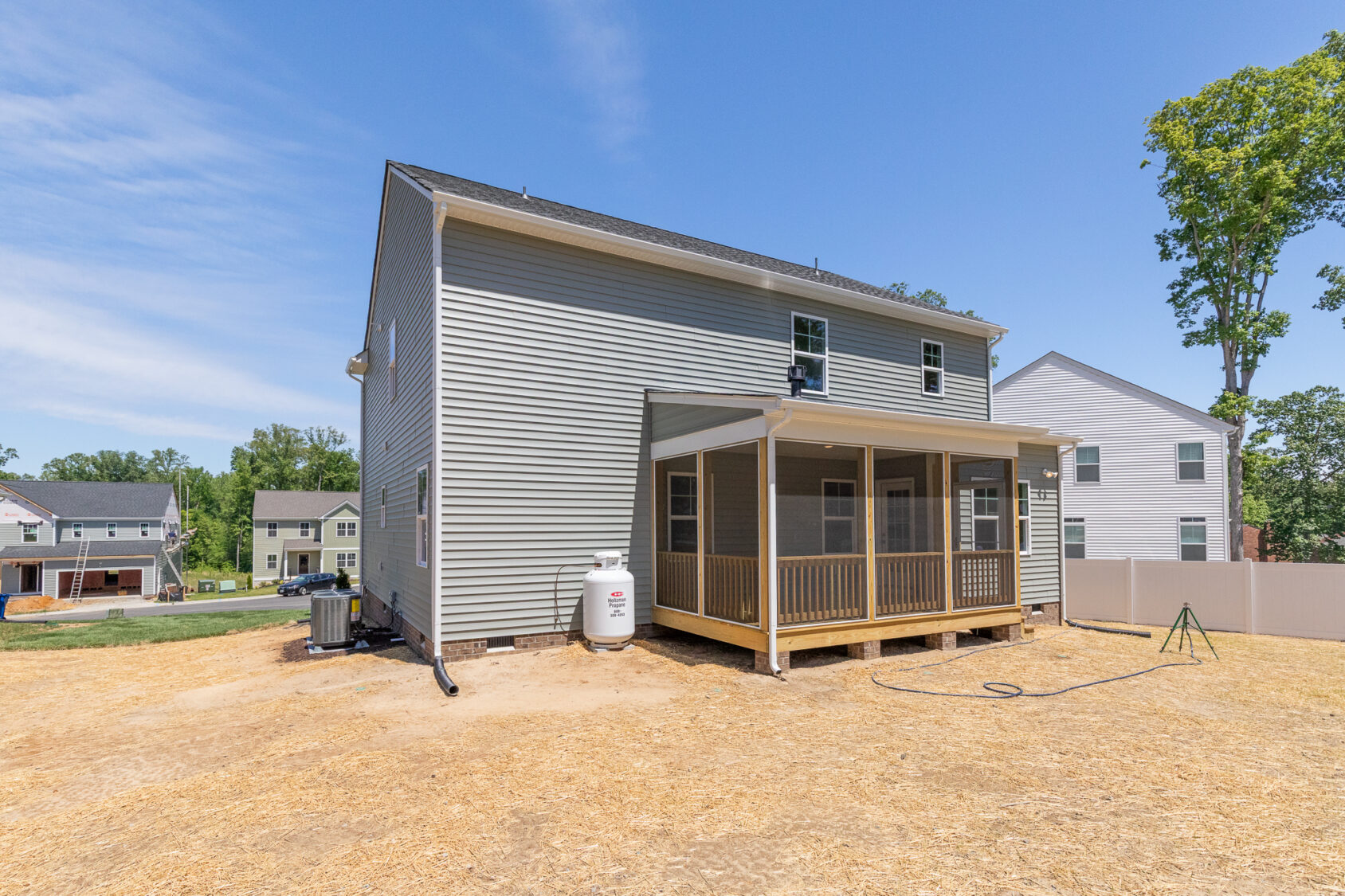 Back view of Poplar Village Lot 26: gray house with screened porch, propane tank, and dry yard under a clear blue sky.