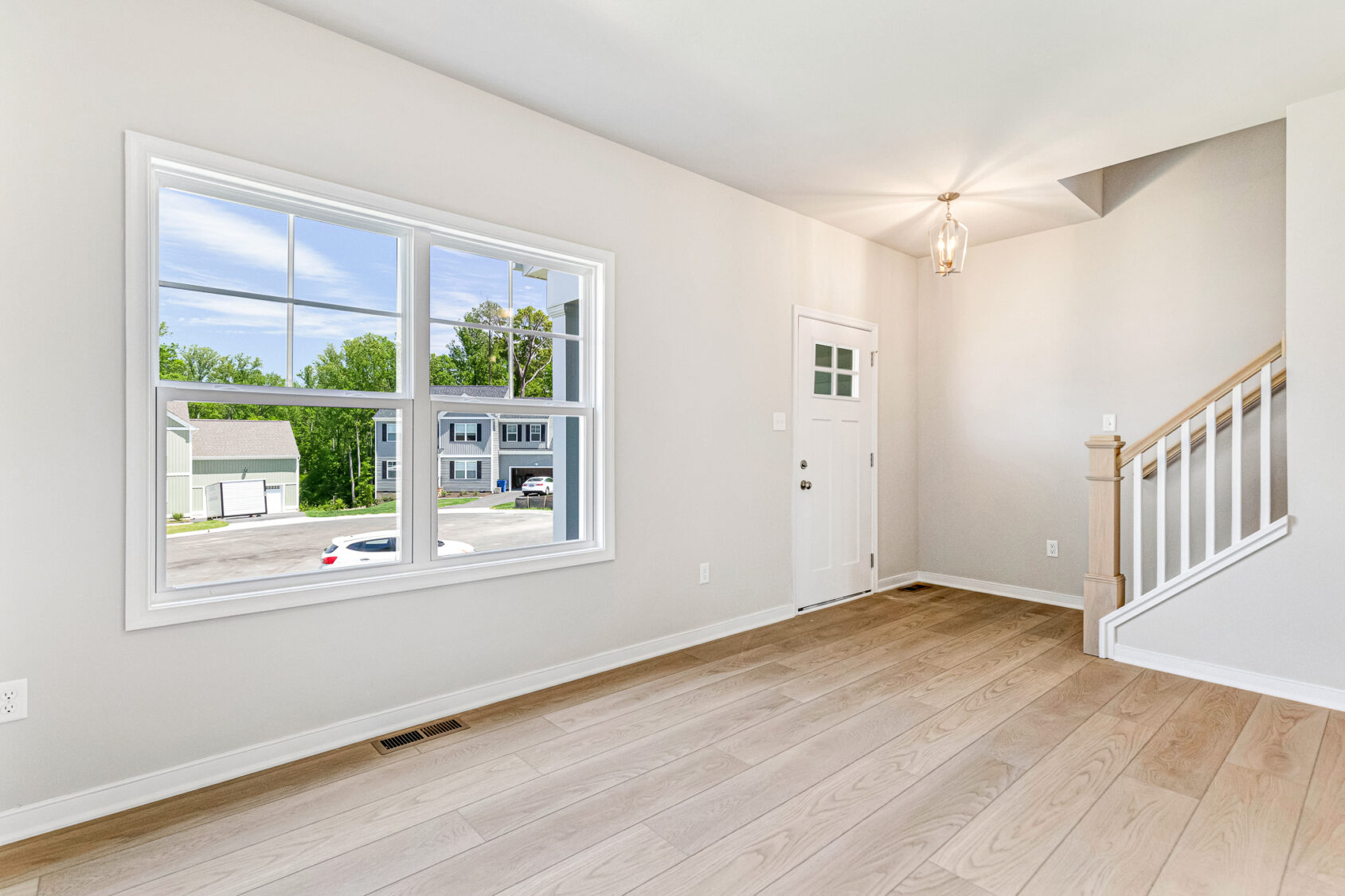Bright, empty room in Poplar Village with light wood floors, large window, stairway, and white front door.