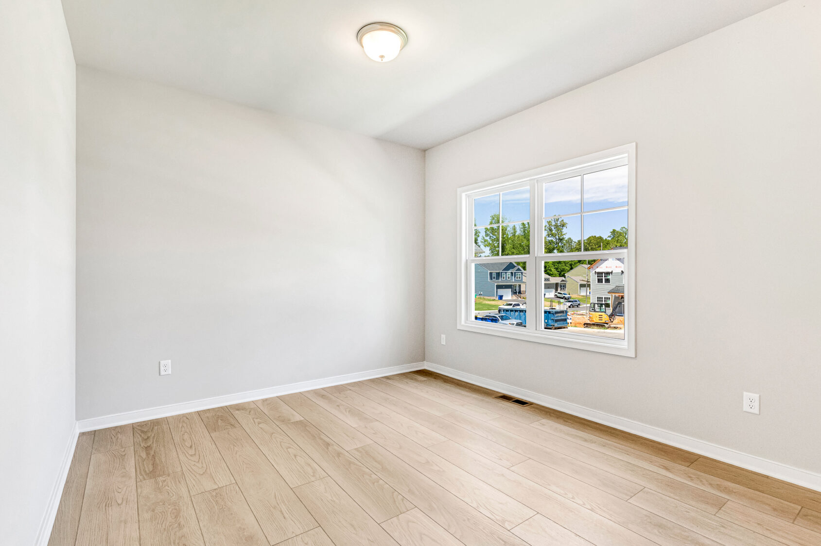 Empty, bright room in Poplar Village with light wood floors, white walls, and a window overlooking Lot 26.