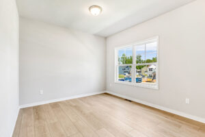 Empty, bright room in Poplar Village with light wood floors, white walls, and a window overlooking Lot 26.