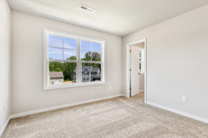 Empty, carpeted bedroom in Poplar Village with a large window, beige walls, and open door to adjacent room.