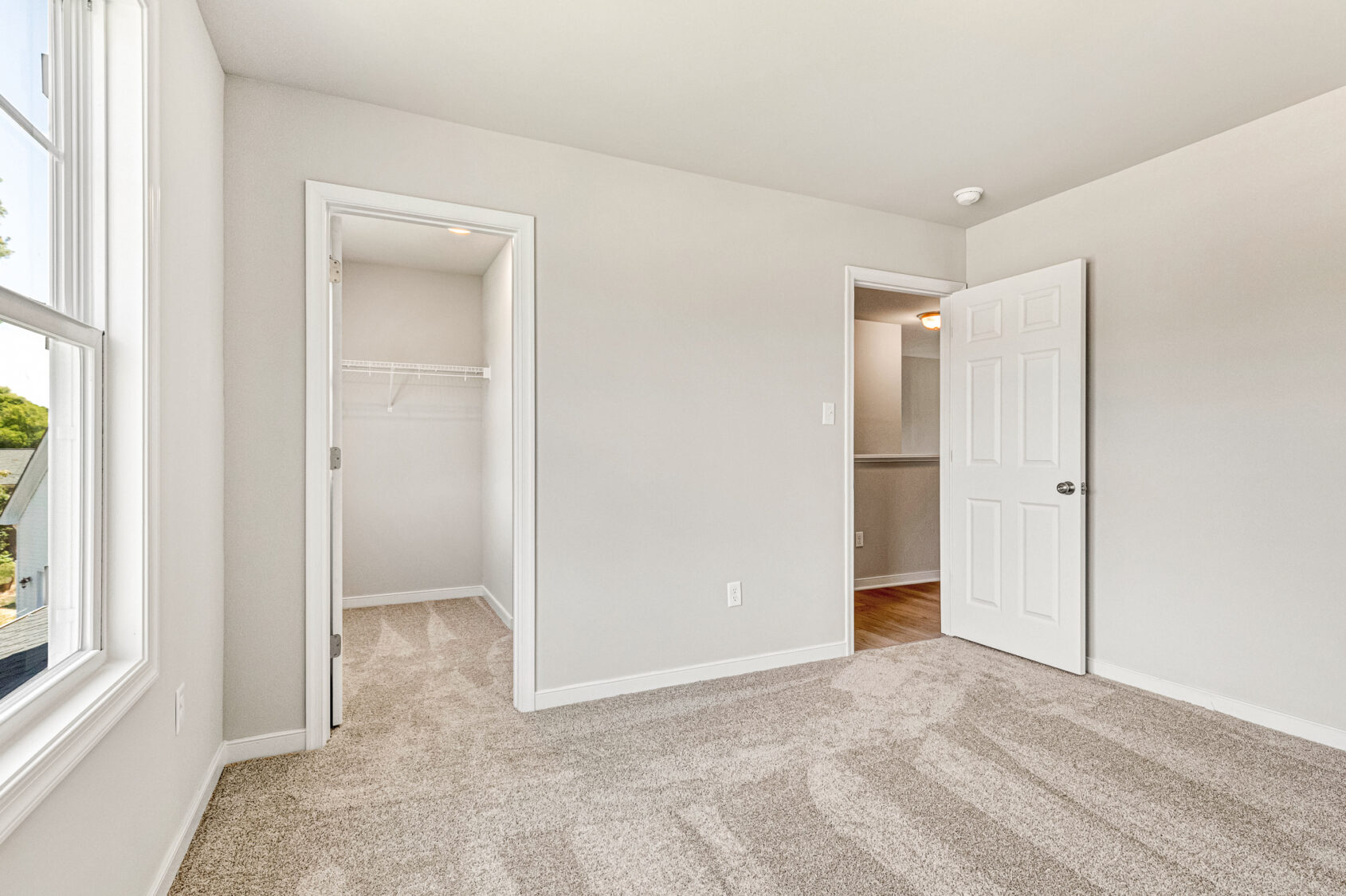 Bright, empty bedroom in Poplar Village's Lot 26 with beige carpet, open closet, and white walls, viewed from a window.