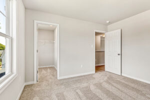 Bright, empty bedroom in Poplar Village's Lot 26 with beige carpet, open closet, and white walls, viewed from a window.