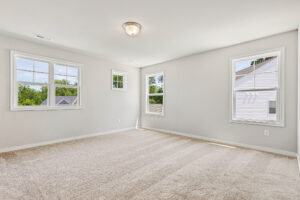 Bright, empty carpeted room in Poplar Village, Lot 26, with three windows, white walls, and a ceiling light fixture.