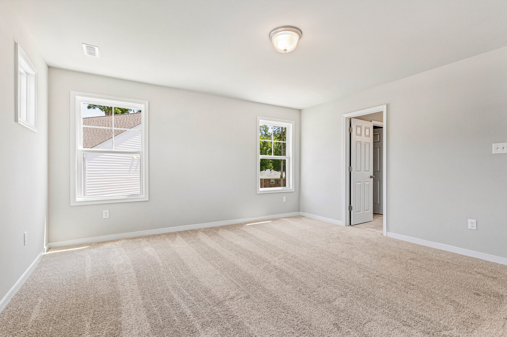 Bright, empty carpeted room in Poplar Village with white walls, two windows, ceiling light, and open door.