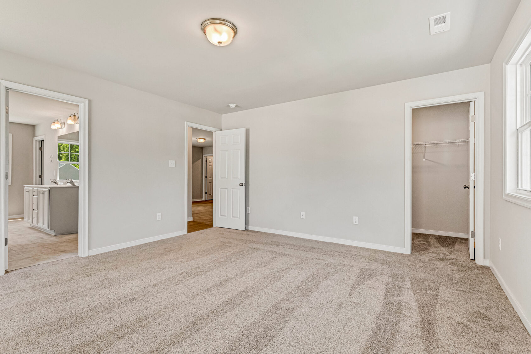 Empty, carpeted bedroom in Poplar Village Lot 26 with beige walls, large window, and open doorways to bath, closet, and hall.