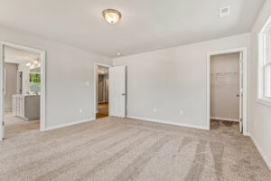 Empty, carpeted bedroom in Poplar Village Lot 26 with beige walls, large window, and open doorways to bath, closet, and hall.
