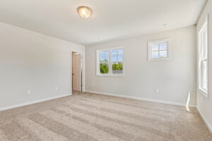 Empty, carpeted room in Poplar Village Lot 26 with beige walls, two windows, and a door to an adjoining space under ceiling light.