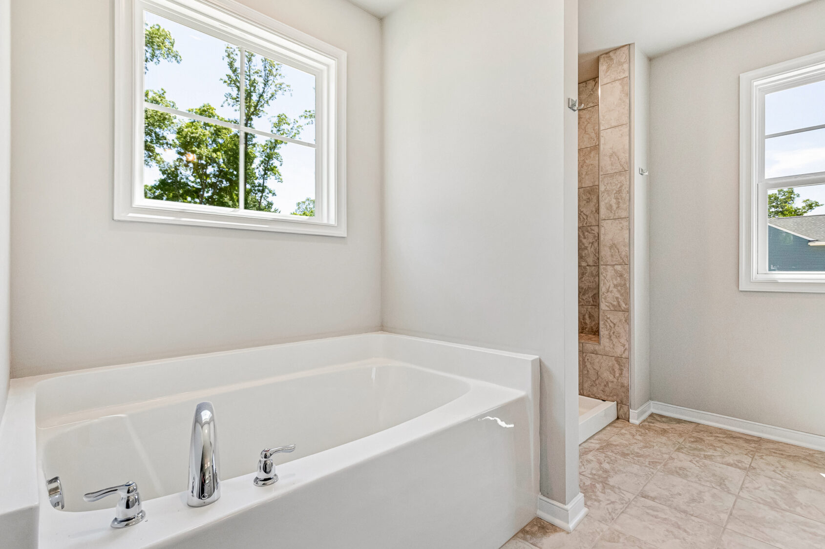 Bright bathroom in Poplar Village with a white bathtub, tiled floor, walk-in shower, and two windows with outdoor views.