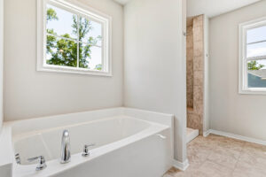 Bright bathroom in Poplar Village with a white bathtub, tiled floor, walk-in shower, and two windows with outdoor views.