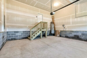 Unfinished garage interior in Poplar Village, Lot 26, with concrete floor, exposed drywall, wooden stairs, and water heater.