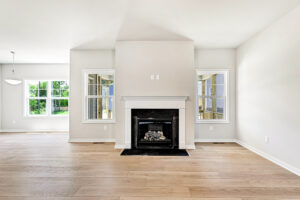 Bright empty living room in Poplar Village with light wood floors, fireplace, and two windows on Lot 26.