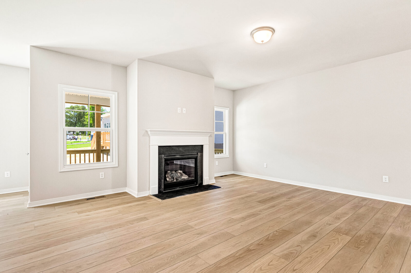 Bright, empty living room in Poplar Village Lot 26 with light wood floors, white walls, fireplace, and two windows.