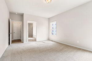 Empty, bright Somerset Hill bedroom with beige carpet, white walls, ceiling light, window, and two open doors to other rooms.