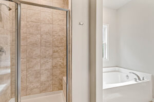 Glass shower with beige tiles, Lot 8 Somerset Hill, next to a white bathtub under a window in a bright bathroom.