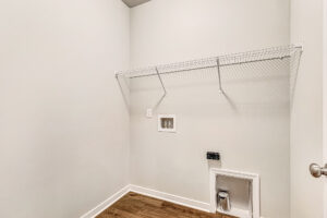 Empty laundry room with white walls, shelf, and hookups on wood floor in Somerset Hill’s Lot 8.