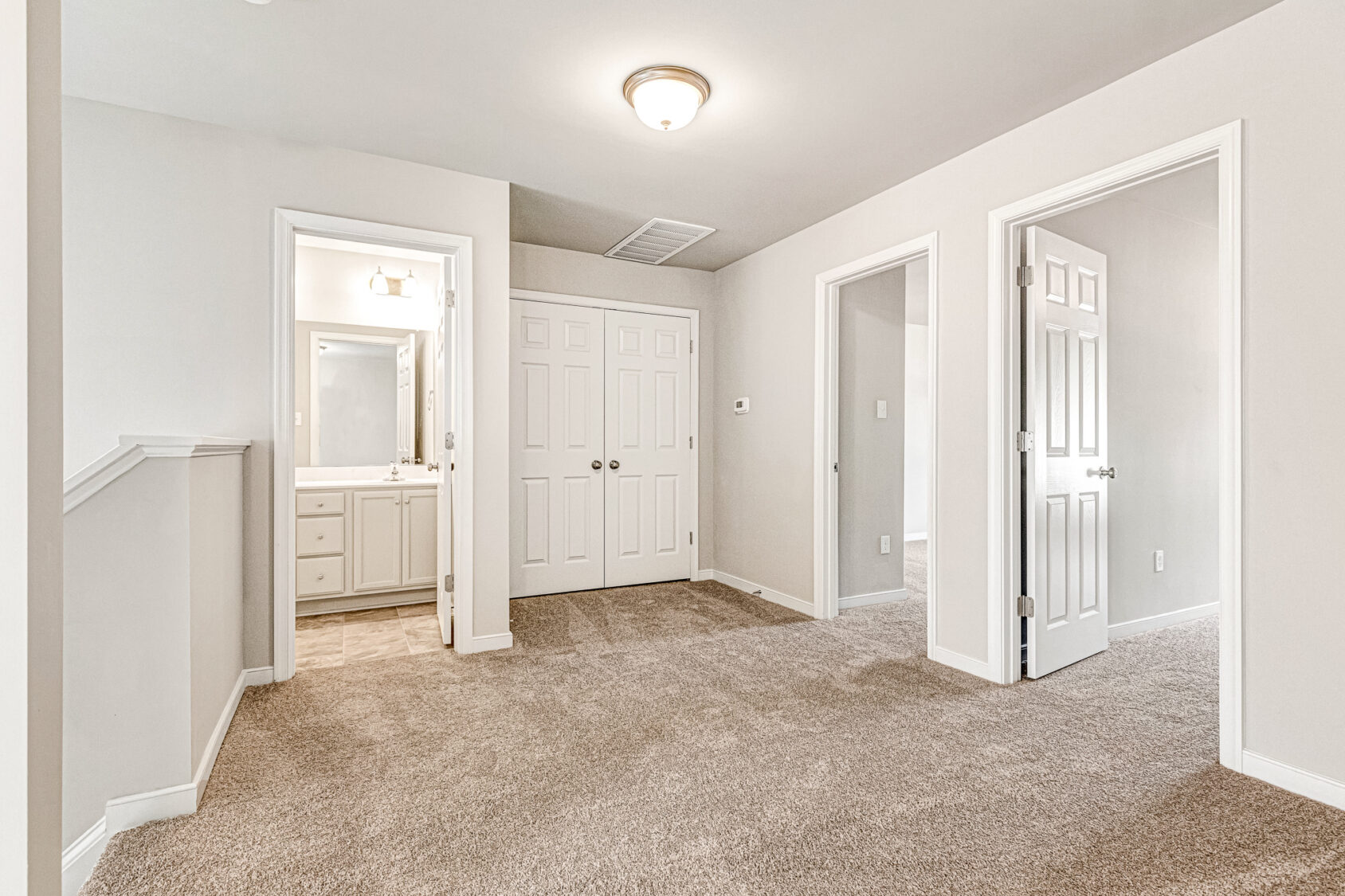 Bright, empty carpeted room in Somerset Hill with three white doors, one opening to a bath with large mirror and vanity.