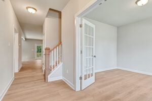 Bright hallway in Poplar Village Lot 7 with light wood floors, staircase, and French doors to an empty white-walled room.