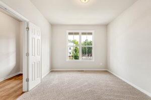 Bright, empty Somerset Hill room with beige carpet, white walls, large window, and open door to Lot 8’s wood-floored area.