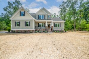 Two-story beige house in Shirley Commons, Lot 7, with a porch, stone accents, and a spacious front yard surrounded by trees.