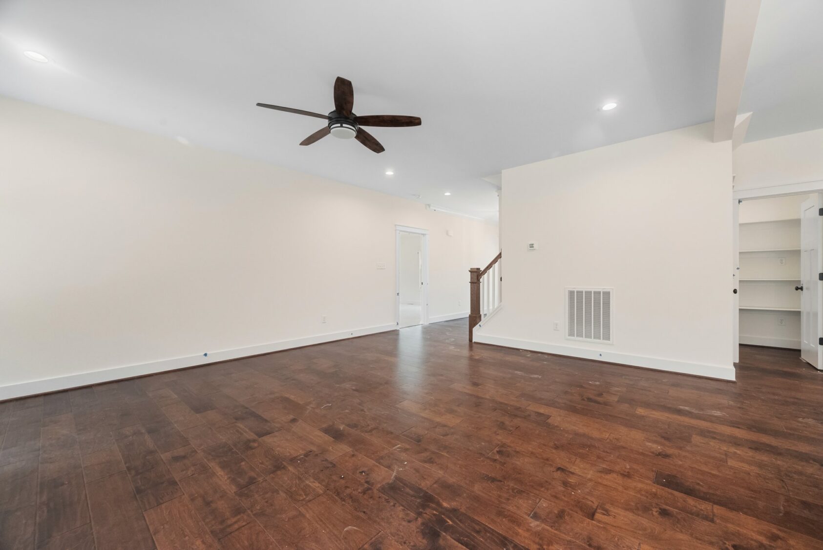 Spacious empty living room with dark wood floors in Shirley Commons Lot 7, white walls, ceiling fan, and open stairs.