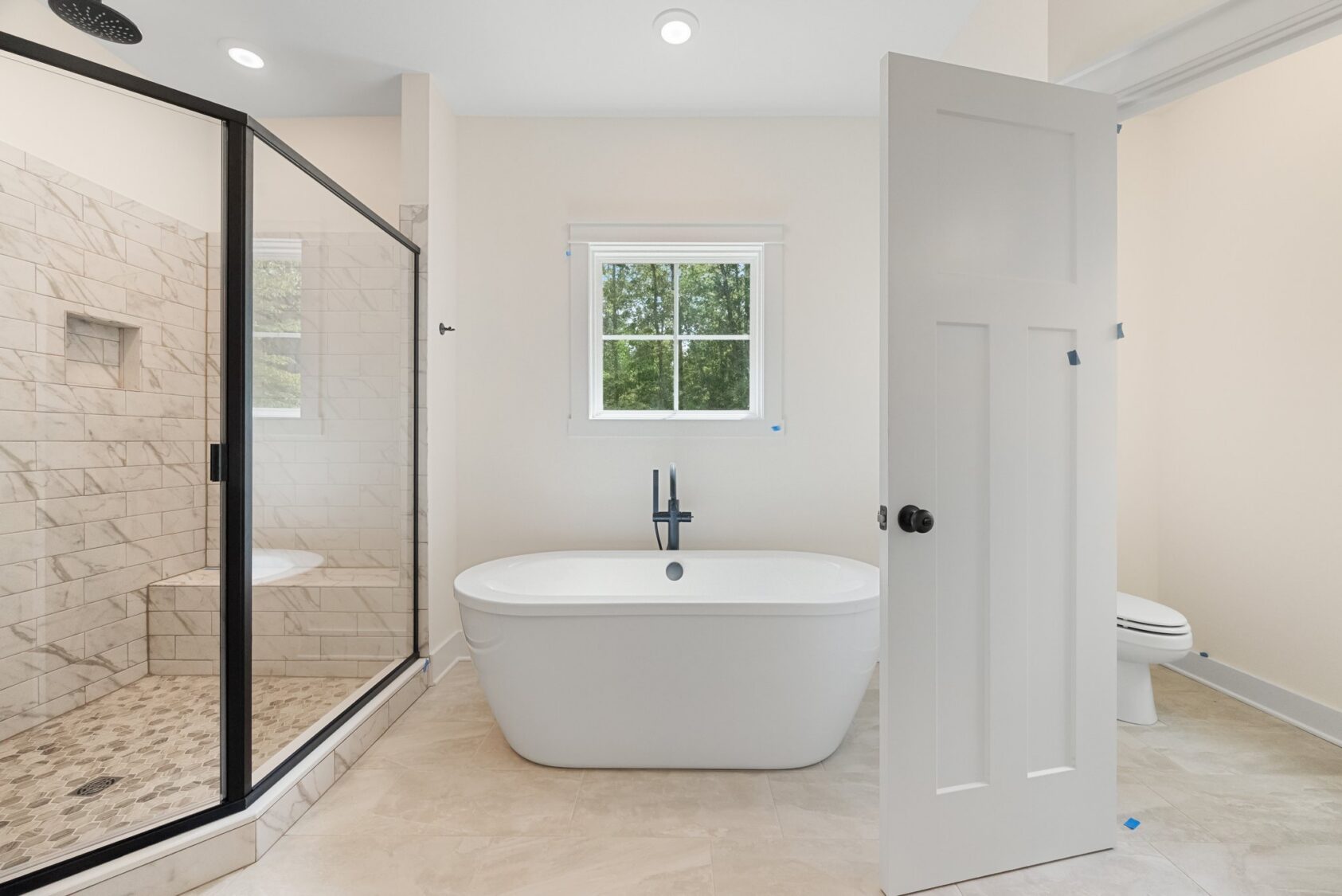 Modern bathroom in Shirley Commons Lot 7 with a freestanding tub, glass shower, window, and neutral tile walls and floor.
