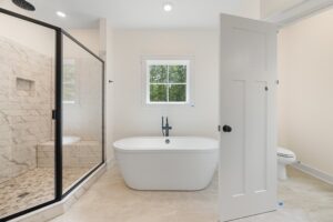 Modern bathroom in Shirley Commons Lot 7 with a freestanding tub, glass shower, window, and neutral tile walls and floor.