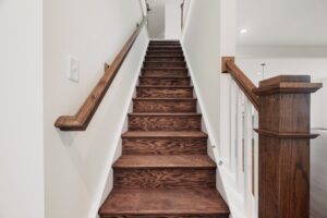 A staircase with dark wooden steps and a wooden handrail leads upward in Lot 7 at Shirley Commons.