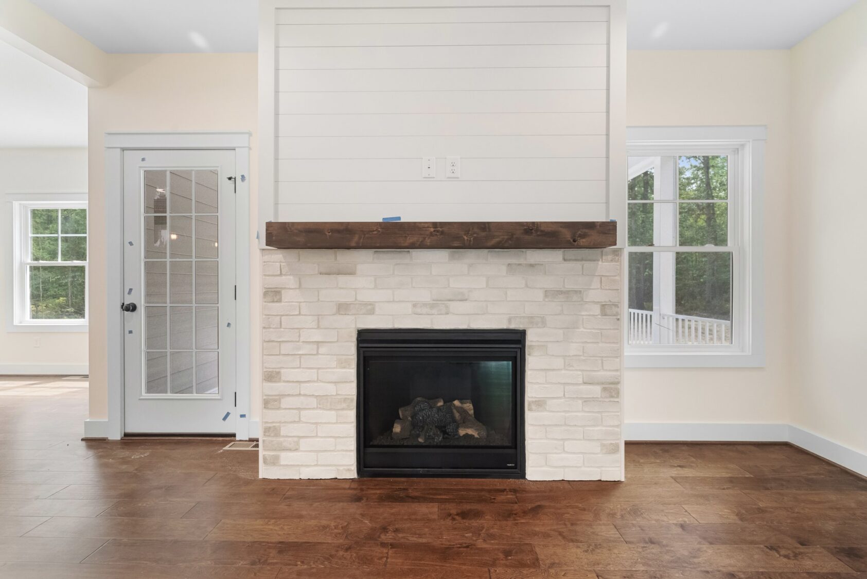 White brick fireplace with wooden mantel in bright Shirley Commons room, near windows and glass-paned door on Lot 7.