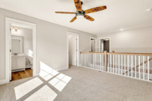 Sunlit carpeted room at Somerset Hill, with ceiling fan, white railing, and two doors, one leading to a bathroom.