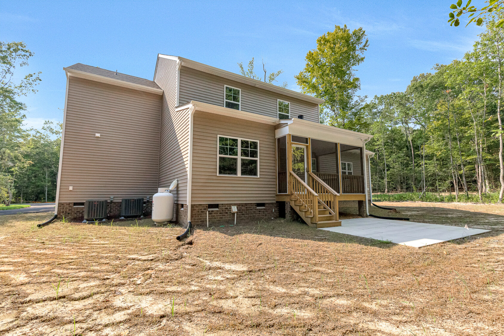 Two-story tan house on Lot 7, Somerset Hill, with a small porch, patio, and forested backdrop under a clear blue sky.