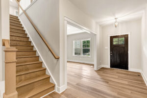 Bright Somerset Hill entryway with wooden stairs, light floors, and a dark front door next to a sunny room. Lot 7.
