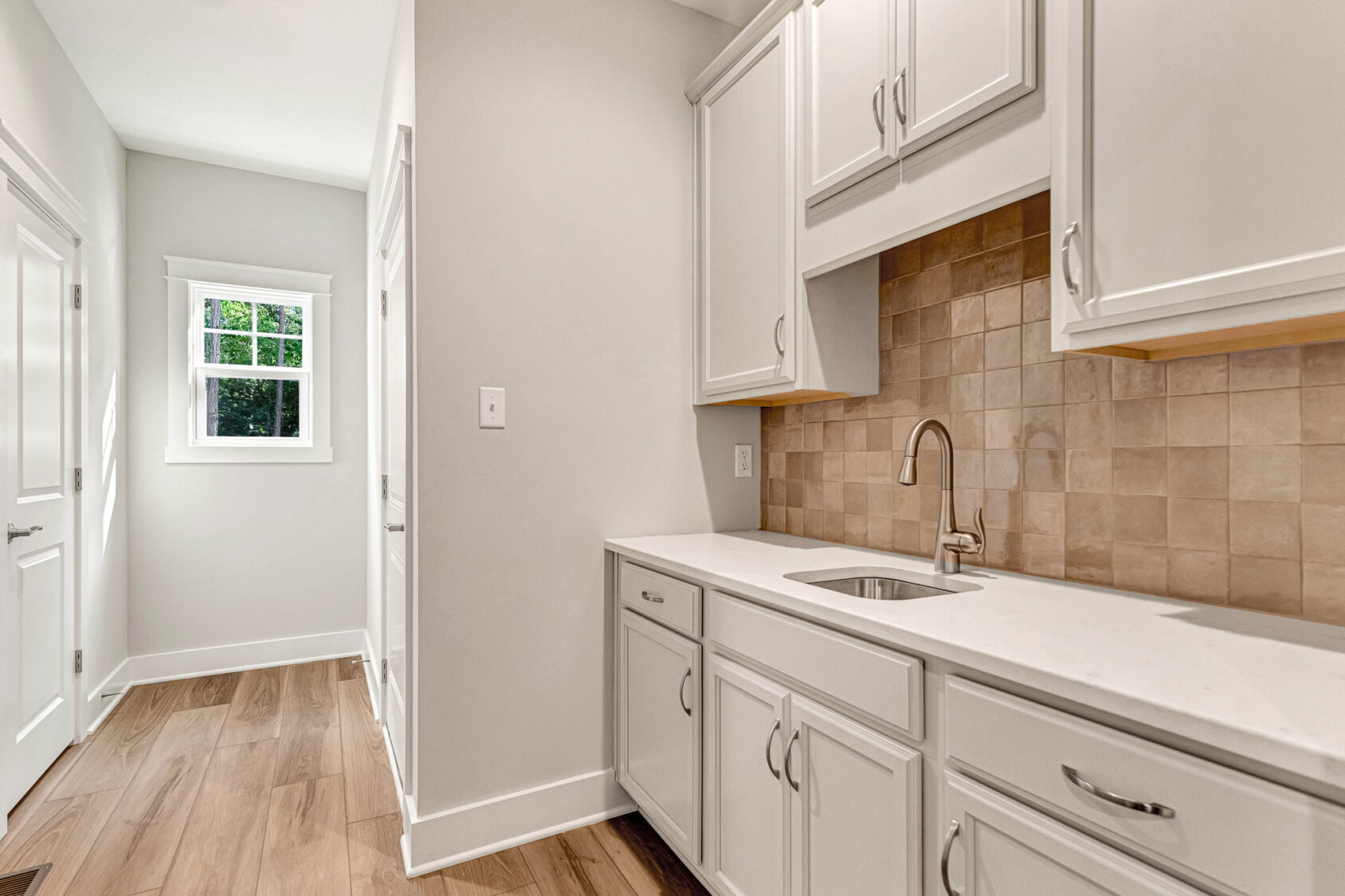Modern laundry room in Somerset Hill Lot 7 with white cabinets, tan tile backsplash, wood floor, and natural window light.