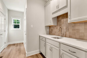 Modern laundry room in Somerset Hill Lot 7 with white cabinets, tan tile backsplash, wood floor, and natural window light.