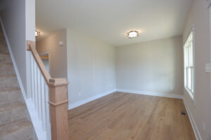 Empty living room with wood floors in Somerset Hill, beige walls, a window, and carpeted stairs with white railing on left.