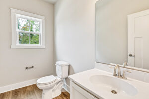 Bright Somerset Hill bathroom with white sink, toilet, mirror, and window overlooking green trees; wood floor visible.